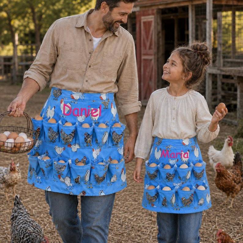 May include: Two blue egg-collecting aprons featuring a repeating chicken pattern and embroidered names. The larger apron displays the name Daniel in pink, while the smaller apron features the name Maria in pink, both designed with multiple pockets.