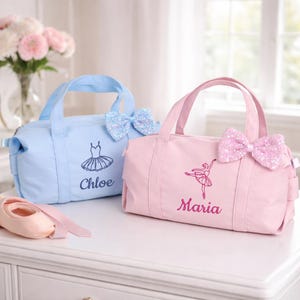 May include: Two ballet-themed duffel bags, one light blue and one pink, sit on a white surface. Each bag has a matching sequined bow and embroidered ballet-themed design with a name. A ballet shoe and flowers are in the background.