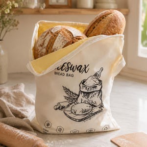 May include: A cream-coloured beeswax bread bag with a yellow interior lining, containing two round loaves of crusty bread. The bag features black printed illustrations of a flour sack, wheat, a rolling pin, and the text Beeswax Bread Bag.