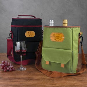May include: Two wine bottle carrying bags. One is black with a red trim and strap, the other is green with a brown strap. Both have a leather-like patch with text. A wine glass with red wine and a bunch of grapes are in the foreground.