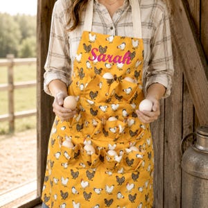 May include: A mustard yellow apron featuring a repeating pattern of chickens and roosters, with multiple pockets designed for holding eggs. The name Sarah is embroidered in bright pink script across the upper chest area of the apron.