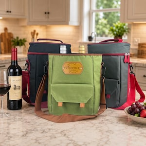 May include: Three insulated wine bottle carriers in navy, green, and gray with red and brown accents. The green carrier has a front pocket and a leather patch with the text "Phoenix". A bottle of red wine and a wine glass are on the left.