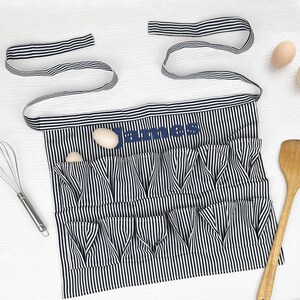 May include: A black and white vertically striped apron featuring two rows of gathered pockets and long waist ties. The name James is embroidered in dark blue thread across the upper section. The apron is designed for holding eggs.