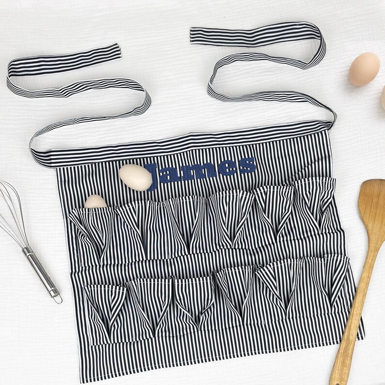 May include: A black and white vertically striped apron featuring two rows of gathered pockets and long waist ties. The name James is embroidered in dark blue thread across the upper section. The apron is designed for holding eggs.