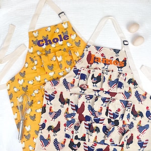 May include: Two aprons with multiple pockets and neck straps. One is yellow with a white chicken pattern and the name Chole in purple. The other is cream with a rooster pattern featuring American flag motifs and the name James in orange.