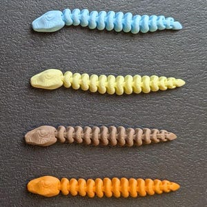 May include: A collection of seven articulated snake toys in various colors, including white, green, blue, yellow, brown, orange, and pink. Each snake has a detailed head and a segmented body, all arranged on a dark surface.