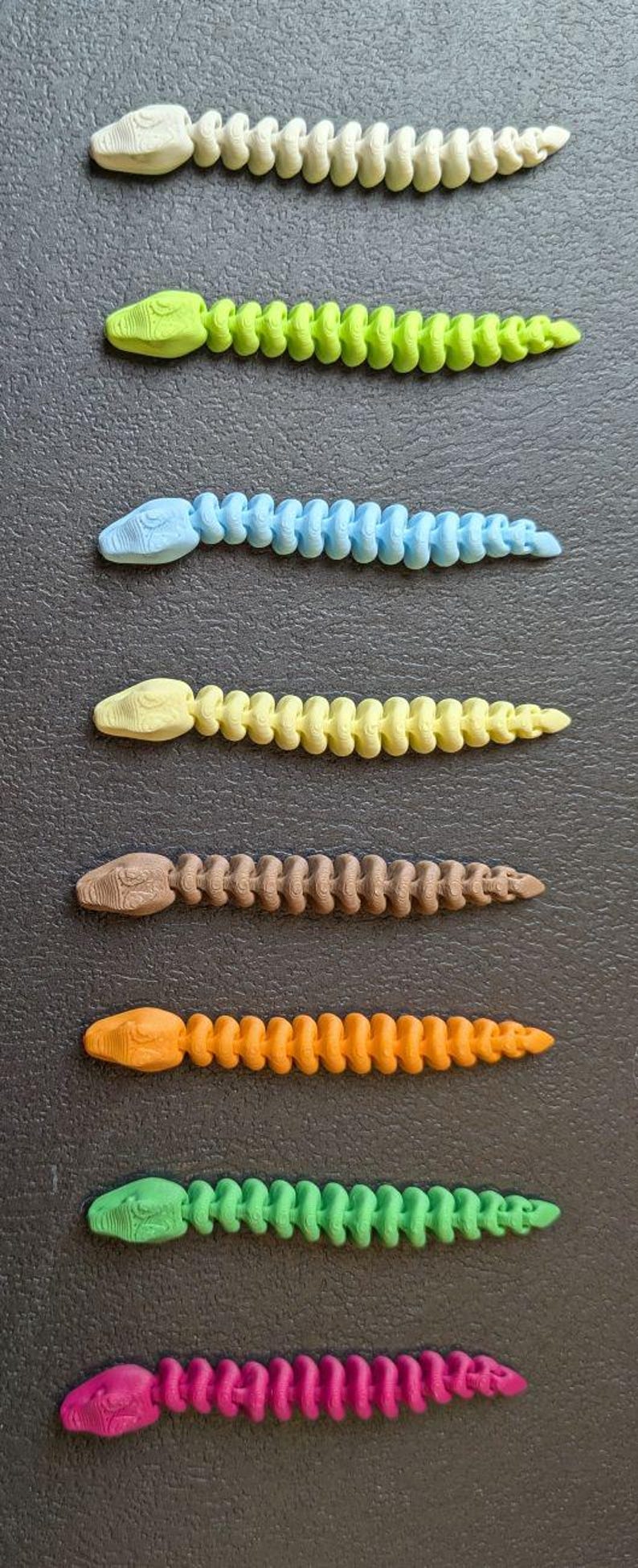 May include: A collection of seven articulated snake toys in various colors, including white, green, blue, yellow, brown, orange, and pink. Each snake has a detailed head and a segmented body, all arranged on a dark surface.