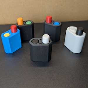 May include: A collection of small, hexagonal-shaped devices in various colors. Each device has two circular openings and a cylindrical button on top. Colors include black, blue, white, and orange, with buttons in red, yellow, green, and black.