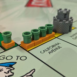 May include: Monopoly board game with custom green pipe and gray castle game pieces. The board shows property names like California Avenue and Pacific Avenue. The game pieces are on a light green board.