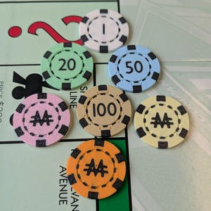 May include: A collection of colorful game chips with values of 1, 20, 50, and 100, along with chips featuring a double "M" symbol. The chips are arranged on a Monopoly game board, with the word "AVENUE" visible.