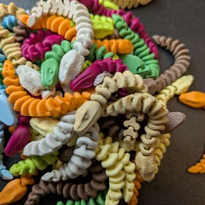 May include: A collection of colorful, 3D-printed snake toys in various colors, including orange, green, blue, and white. The snakes are coiled and have detailed heads. The toys are arranged in a pile, with one snake partially separated from the group.