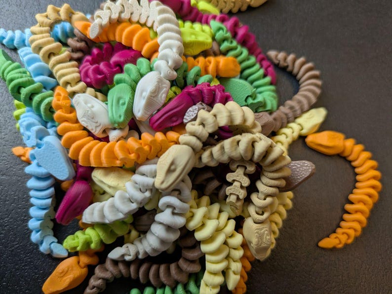 May include: A collection of colorful, 3D-printed snake toys in various colors, including orange, green, blue, and white. The snakes are coiled and have detailed heads. The toys are arranged in a pile, with one snake partially separated from the group.