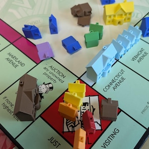 Puede incluir: Primer plano de un tablero de juego de Monopoly con casas y hoteles en varios colores, incluyendo azul, amarillo, rojo y marr&oacute;n. El tablero es verde y tiene las palabras "Maryland Avenue", "Connecticut Avenue", "Vermont Avenue", "Auction", "Just", "Visiting" y "Price 6" impresas en &eacute;l.