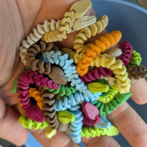 May include: A handful of colorful, 3D-printed snake toys in various colors, including beige, brown, orange, yellow, green, blue, and pink. The toys are detailed with segmented bodies and snake head designs. The toys are held in a hand.