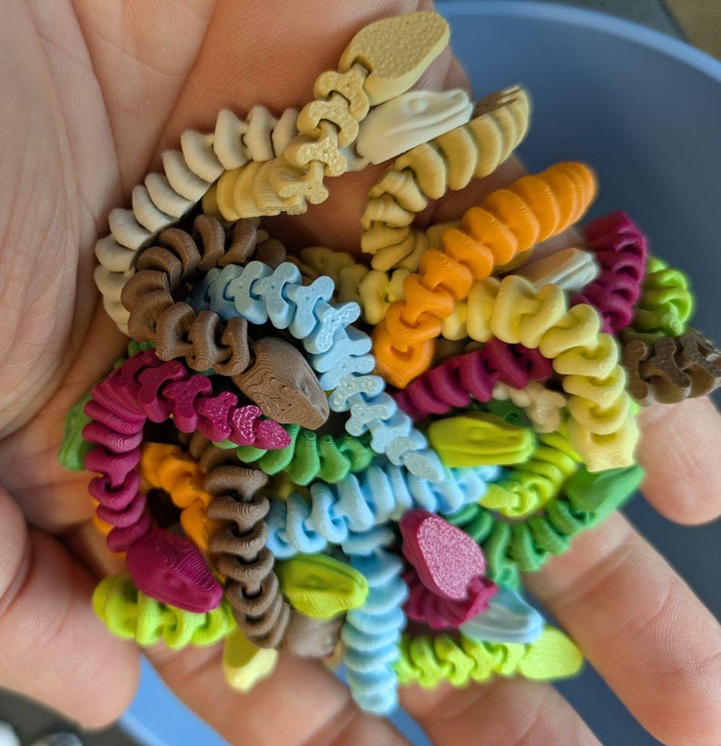 May include: A handful of colorful, 3D-printed snake toys in various colors, including beige, brown, orange, yellow, green, blue, and pink. The toys are detailed with segmented bodies and snake head designs. The toys are held in a hand.