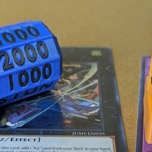 May include: Blue and orange game dice with black text indicating numbers and "STUN". The dice are placed on top of trading cards. The blue die shows the numbers 1000, 2000, and 3000. The orange die shows the numbers 1000, 2000, and 3000.