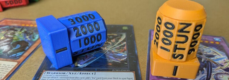 May include: Blue and orange game dice with black text indicating numbers and "STUN". The dice are placed on top of trading cards. The blue die shows the numbers 1000, 2000, and 3000. The orange die shows the numbers 1000, 2000, and 3000.
