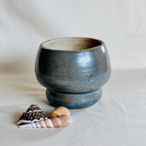 Puede incluir: Un cuenco de cerámica redondeado con un esmalte azul grisáceo moteado y un borde ancho y acampanado. El cuenco presenta una cresta horizontal distintiva cerca de la base. Junto al cuenco hay tres conchas marinas, incluida una concha oscura con patrones y dos conchas en espiral alargadas de color claro.