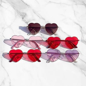 Puede incluir: Colección de gafas de sol con forma de corazón en varios colores, incluyendo rojo, rosa, morado y marrón. Las gafas tienen monturas metálicas y lentes tintadas. Están dispuestas sobre una superficie de mármol blanco.