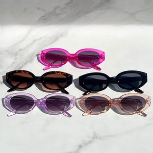 Puede incluir: Seis pares de gafas de sol ovaladas con diferentes colores de montura y lentes. Las monturas son rosa, marrón, negro, morado y marrón claro. Las lentes son oscuras.