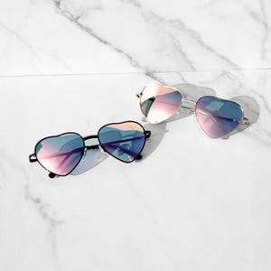 Peut inclure: Deux paires de lunettes de soleil en forme de cœur avec des verres irisés. Une paire a une monture noire et l'autre une monture dorée. Les verres reflètent des nuances de rose, de bleu et de violet. Les lunettes sont présentées sur une surface en marbre blanc.