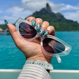 Puede incluir: Gafas de sol blancas de ojo de gato con lentes oscuros. Las gafas se sostienen en una mano con esmalte de uñas de brillo turquesa y plateado.