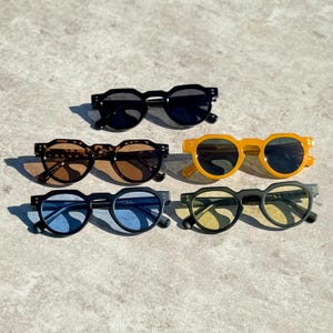 May include: Six pairs of sunglasses in various colors are displayed on a light gray surface. The sunglasses include black, tortoise shell, yellow, blue, and green frames. Each pair has dark lenses and a round shape.