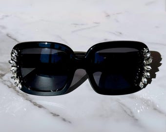 May include: Black square sunglasses with rhinestones on the sides. The sunglasses have dark lenses.