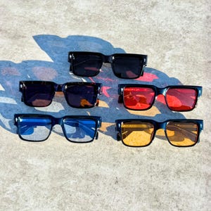 May include: Six pairs of sunglasses with black frames and various lens colors, including black, red, blue, and yellow. The sunglasses are arranged on a concrete surface, showcasing their different styles.