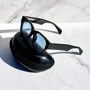 Puede incluir: Gafas de sol negras con lentes azules sobre un estuche negro texturizado. Las gafas de sol tienen un diseño de montura geométrica. El estuche tiene cierre de cremallera. El fondo es una superficie de mármol blanco.