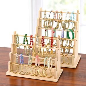 Op de afbeelding: Twee houten sieradenstandaards met een verscheidenheid aan armbanden. De armbanden omvatten gouden armbanden en kralenmodellen in verschillende kleuren zoals groen, rood en roze. De standaards zijn gemaakt van lichtgekleurd hout.