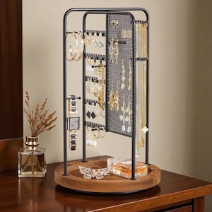 Op de afbeelding: Een zwarte metalen sieradenorganizer met een houten basis. De organizer heeft een gaaspaneel voor het ophangen van kettingen en oorbellen, en een stang voor armbanden. Een horloge en diverse sieraden worden getoond. Een parfumflesje en decoratieve vlinder staan op de basis.