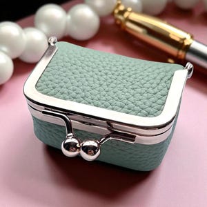 以下が含まれることがあります： テクスチャード加工された革のような外観の、小さな長方形のセージグリーンの小銭入れ。シルバーカラーの金属フレームと、2つの球形アクセントが付いたクラスプクロージャーが特徴です。小銭入れはソフトピンクの背景に配置されています。