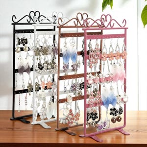 Peut inclure: Trois présentoirs à boucles d'oreilles en métal noir, blanc et rose, chacun contenant de nombreuses paires de boucles d'oreilles. Les présentoirs ont un design supérieur décoratif et sont remplis de différents styles de boucles d'oreilles.