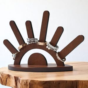May include: A dark brown wooden ring holder with a semi-circular base and five upright, angled posts. Several rings of various designs are displayed on the posts. The holder sits on a wooden surface against a white background.
