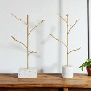 May include: Two gold-coloured metal jewellery trees with a branch design. One tree has a square, speckled white base, and the other has a round base. Both are displayed on a wooden surface.