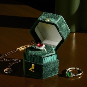 May include: A dark green velvet ring box with a gold clasp, open to reveal a silver ring with a red gemstone. A gold necklace and a silver ring with a green gemstone are also visible.