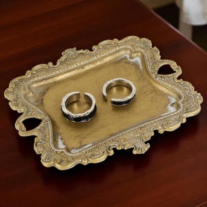 Puede incluir: Dos anillos plateados con detalles negros descansan sobre una bandeja dorada ornamentada. Los anillos tienen un diseño abierto. La bandeja tiene bordes y asas decorativas, colocada sobre una superficie de madera oscura.