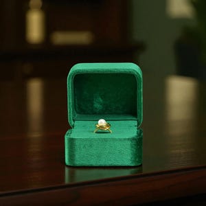 Puede incluir: Un anillo de oro con una perla blanca en el centro se encuentra dentro de un joyero de terciopelo verde esmeralda abierto. El joyero es rectangular con una tapa con bisagras. El fondo es un interior oscuro y borroso.