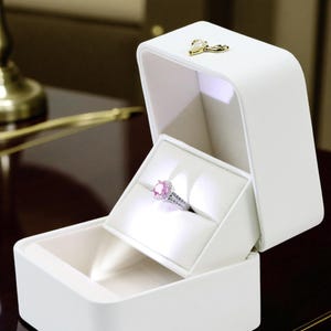 May include: A white ring box with a hinged lid, open to reveal a pink gemstone ring. The ring is set in a silver band and surrounded by smaller stones. The box has a gold-colored clasp.