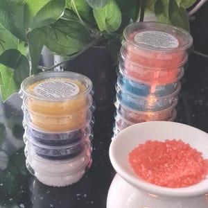 May include: A selection of colourful bath salts in clear plastic containers. The salts are in shades of yellow, blue, and white. A white ceramic oil burner holds red bath salts. Green foliage in the background.