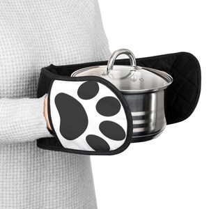 Puede incluir: Manoplas para horno con estampado de huellas de patas en blanco y negro que sostienen una olla de acero inoxidable con tapa de cristal. Las manoplas tienen un reverso negro acolchado y un frente blanco con un diseño de huella de pata negra.