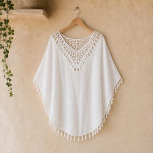 May include: A white poncho featuring a V-shaped neckline with intricate crochet detailing. The garment has a loose, flowing silhouette and is finished with a decorative fringe trim along the bottom hem, hanging from a wooden hanger.