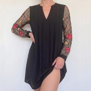 May include: Black dress with sheer sleeves embroidered with red and yellow roses. The dress has a V-neck and pleats down the front. The sleeves are long and fitted, and the dress falls to mid-thigh.