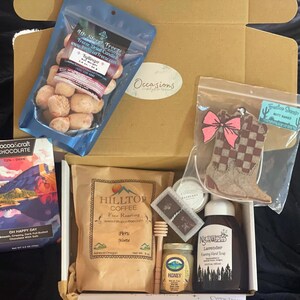Peut inclure: Une boîte en carton marron remplie de divers cadeaux, dont un sac de bonbons lyophilisés, un sac de café, une barre de chocolat noir, une bouteille de miel, une bouteille de savon moussant pour les mains à la lavande, un petit pot de miel, un bâtonnet en bois pour le miel, un petit sac de chocolat et un désodorisant en forme de botte de cowboy en feutre.