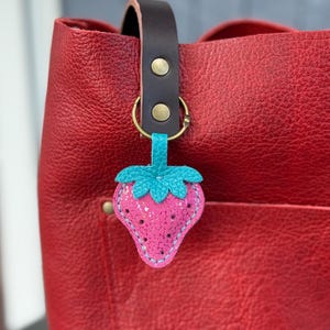 Peut inclure: Un porte-clés fraise rose avec un dessus vert turquoise est attaché à un sac fourre-tout en cuir rouge. Le porte-clés est en feutre et a une texture pailletée. Le sac a une poignée en cuir marron foncé.