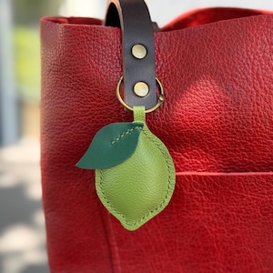 May include: A close-up of a red leather tote bag with a brown leather handle. A green leather lime charm with a green leaf is attached to the handle with a gold ring. The bag has a textured surface.
