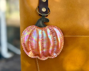 May include: A pumpkin-shaped keychain charm with an iridescent, orange-pink finish. The charm has stitched details and a green stem. It is attached to a brown leather strap with a gold-colored ring.