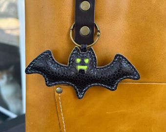 May include: Black leather bat keychain with a green face and eyes. The bat is attached to a brown leather strap with a gold-colored ring. A Halloween-themed accessory.