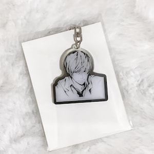 May include: A black and white anime-style keychain featuring a male character with light-colored hair and a serious expression. The keychain is attached to a silver-colored metal clasp and is on a white card.